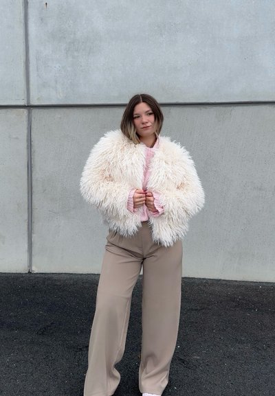 Mujer de pie contra una pared de cemento, vistiendo una chaqueta blanca esponjosa, suéter rosa, pantalones beige de pierna ancha y zapatos blancos.