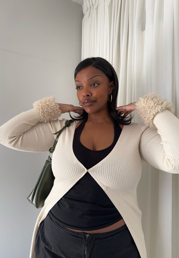 Keskitumman ihonvärin ja mustat hiukset omaava nainen korjaa karvaisten kalteiden neulettaan, pukeutuneena mustaan toppiin ja housuihin, seisten valkoisten verhojen vieressä.