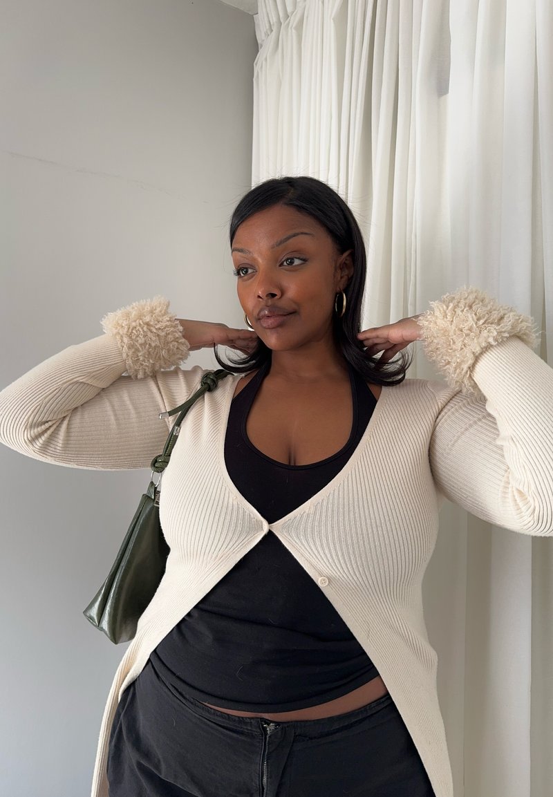 Femme à la peau brun moyen et aux cheveux noirs ajuste son cardigan avec des poignets en fourrure, portant un haut et un pantalon noirs, debout près de rideaux blancs.
