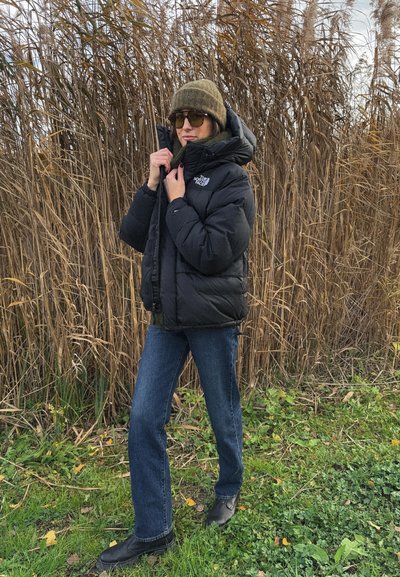 Persona con chaqueta acolchada negra, vaqueros, botas, sombrero y gafas de sol, de pie al aire libre cerca de hierba seca alta con cubierta verde en el suelo.