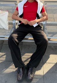 Persona che indossa una maglietta rossa Nike, pantaloni neri e scarpe nere, seduta su una panchina di metallo con una giacca bianca e nera appoggiata sulle spalle.