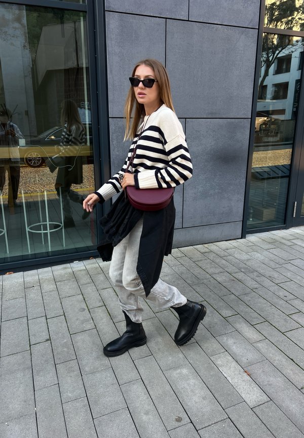 Maglione a righe bianche e nere, jeans grigio chiaro, stivali neri alla caviglia e una borsa a tracolla bordeaux, che cammina su pavimentazione in pietra vicino a una facciata in vetro.