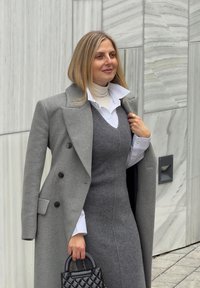 Sivý vlnený kabát cez sivé šaty bez rukávov a bielu košeľu. Čierna prešívaná kabelka. Jemné textúry látok a strihaný dizajn.