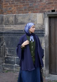 Marineblauwe kimono-jas over een groene gebreide top met grote knopen; blauwe satijnen sjaal; gouden accessoires; spijkerrok; stenen muur op de achtergrond.