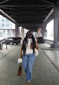 Chaqueta de piel sintética marrón con detalles blancos, vaqueros de mezclilla azul claro y bolso marrón. Gafas de sol y trenzas largas; de pie sobre un pavimento de baldosas.