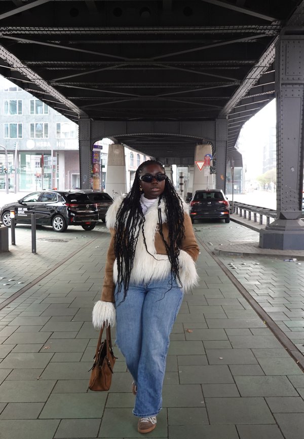 Chaqueta de piel sintética marrón con detalles blancos, vaqueros de mezclilla azul claro y bolso marrón. Gafas de sol y trenzas largas; de pie sobre un pavimento de baldosas.
