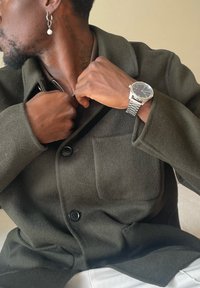 Olívazöld kabátot viselő férfi kigombolja azt, bal csuklóján ezüst óra, fülében gyöngyös karika fülbevaló, balra néz, bézs háttér előtt.