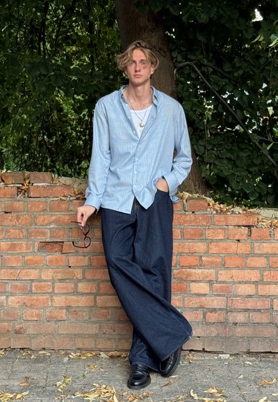 Camisa de cuadros azul claro, jeans de pierna ancha oscuros, zapatos negros. De pie contra una pared de ladrillo con un follaje verde de fondo.