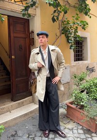 Bež trench kaput preko crnog prsluka i prugastih hlača, smeđe kožne cipele, nosi smeđu torbu na rame, stoji pored zelene biljke.