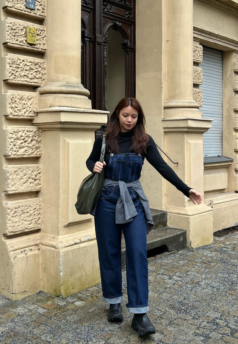 Jeune femme portant un col roulé noir et une salopette en jean marchant sur une rue pavée près d'un bâtiment en pierre beige avec une porte ornée et des colonnes.