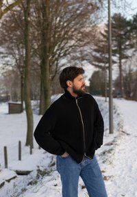 Homme barbu portant un pull noir zippé et un jean bleu, debout les mains dans les poches sur un sentier enneigé près d’arbres et d’un lampadaire.