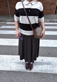 Polo a righe bianco e nero, gonna plissettata scura, borsa a tracolla in pelle marrone con motivo e scarpe marroni con lacci su un attraversamento pedonale.