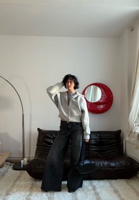 Sweat à capuche gris court avec fermeture éclair, chemise à rayures et jean noir à jambes larges. Sac à main noir. Miroir circulaire rouge sur un mur blanc en arrière-plan. Canapé en cuir marron.