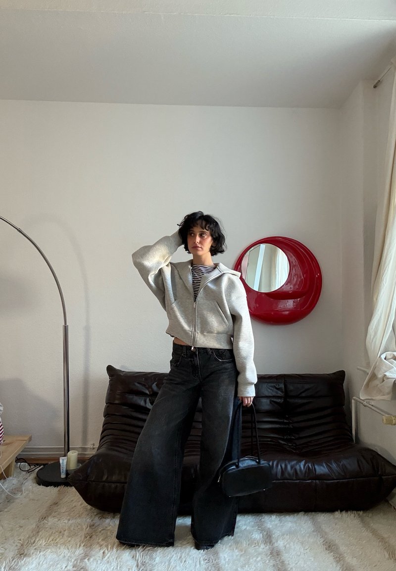 Sweat à capuche gris court avec fermeture éclair, chemise rayée, jean noir pattes larges. Sac à main noir. Miroir circulaire rouge sur un mur blanc derrière. Canapé en cuir marron.