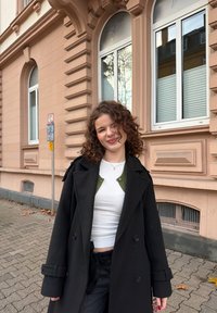 Manteau noir croisé, haut blanc, cardigan vert en dessous. Cheveux bouclés, debout dans une rue pavée près d'un bâtiment beige.