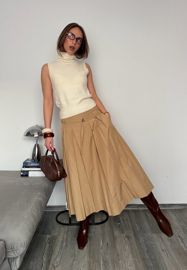 Kvinde iført hvid ærmeløs rullekrave, beige plisseret midinederdel, brune støvler, holder brun håndtaske, stående i rum med lampe og bøger.