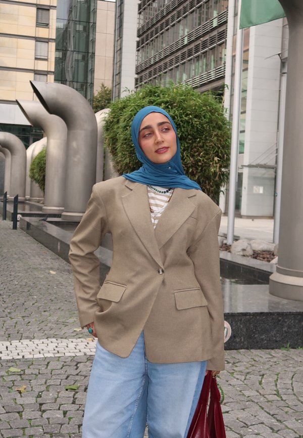 Une femme portant un hijab bleu, un blazer beige et un jean clair pose à côté de sculptures métalliques dans une place urbaine moderne.