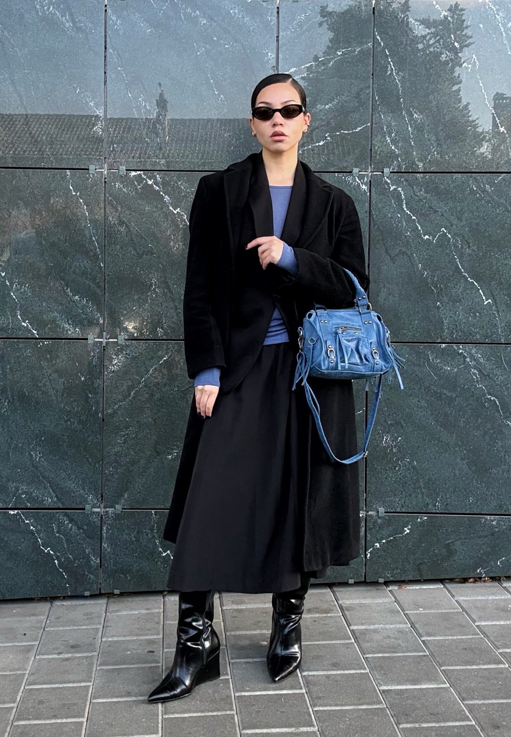 Čierny kabát, modrý top a čierna sukňa v kombinácii so špicatými čiernymi čižmami. Malá modrá kabelka so striebornými kovaniami. Hladké textúry, moderný dizajn.