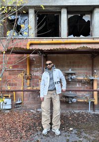 Light gray jacket over a gray hoodie, tan pants, and gray sneakers. Background features red brick, yellow pipes, and broken windows.