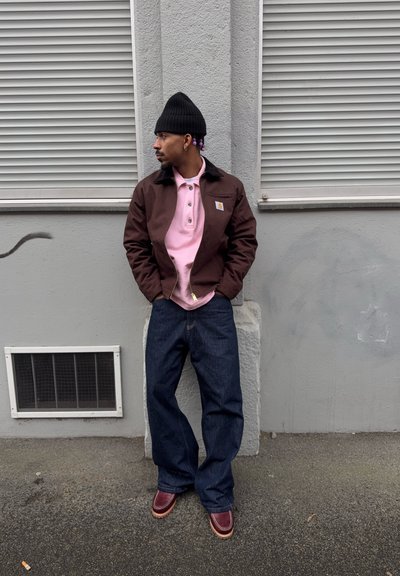 Hombre con gorro negro, chaqueta marrón, camisa rosa, jeans anchos azules y zapatos marrones de pie contra una pared gris con dos persianas cerradas.