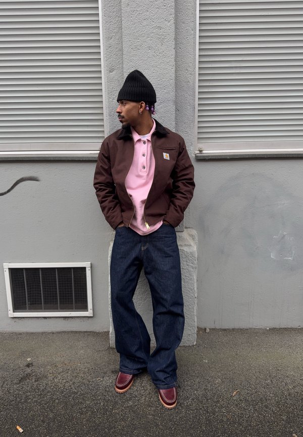 Homme portant un bonnet noir, une veste marron, une chemise rose, un large jean bleu et des chaussures marron, debout contre un mur gris avec deux volets fermés.