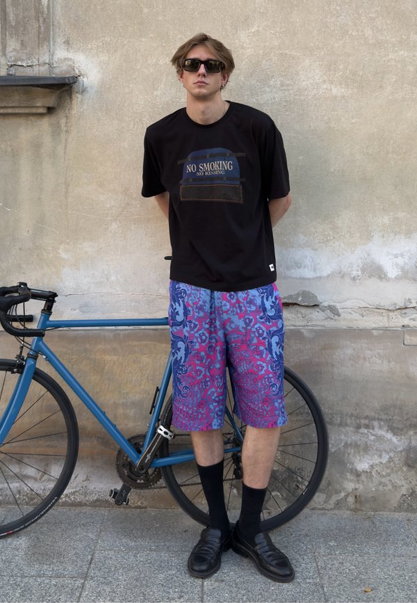 Mladý muž nosiaci slnečné okuliare, čierne tričko s "No Smoking No Kissing" a farebné šortky stojí vedľa modrého bicykla pri stene.