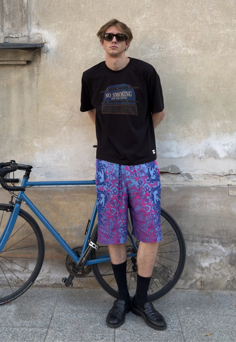 Jeune homme portant des lunettes de soleil, un t-shirt noir avec "No Smoking No Kissing" et un short coloré, debout à côté d'un vélo bleu contre un mur.