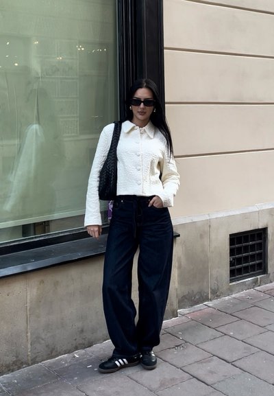 Chaqueta corta blanca de textura con cuello, jeans oscuros de cintura alta y zapatillas negras con rayas blancas. Bolsa negra de tejido.