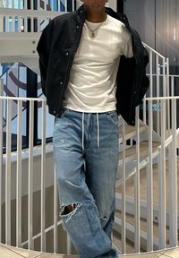 Mann iført svart jakke over hvit t-skjorte, ripped blå jeans med snor i livet, står foran et hvitt spiraltrapprailing innendørs.