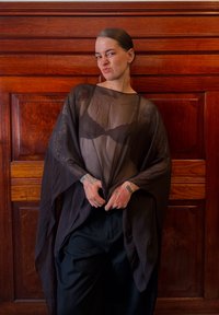 Permatomas juodas kimono stiliaus drabužis su ilgomis rankovėmis, dėvimas ant tamsaus nėriniuoto liemenėlės, derinamas su laisvomis juodomis kelnėmis. Matomi sudėtingi tatuiruotės.