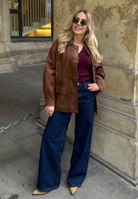 Rjava semiš jakna čez burgundski top, v kombinaciji z širokimi temnimi jeanskimi hlačami in ostrobežnimi bež petami. Kroglaasta sončna očala dopolnjujejo videz.