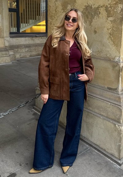 Chaqueta de ante marrón sobre una blusa burdeos, combinada con jeans de mezclilla oscuros de pierna ancha y tacones beiges de punta. Unas gafas de sol circulares completan el look.