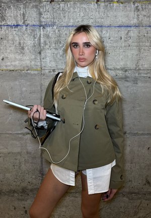 Gabardina verde oliva de doble botonadura con botones grandes, llevada sobre una camisa blanca de cuello y pantalones cortos, sosteniendo un ordenador portátil con auriculares.