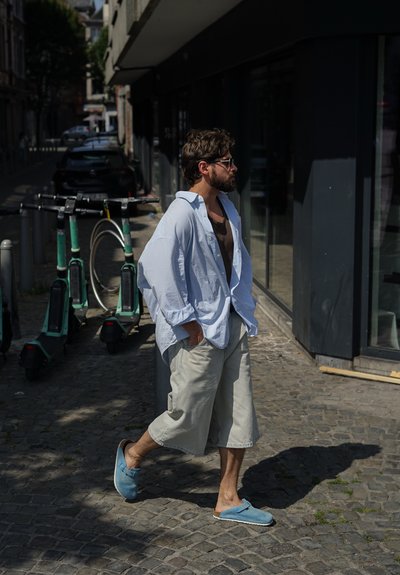 Camisa de botones azul claro de gran tamaño, camiseta marrón debajo, pantalones cortos beige y zapatos de lona azules. Calle de adoquines, patinetes eléctricos al fondo.