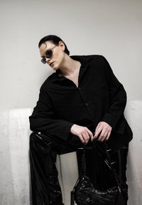 Chemise noire surdimensionnée à boutons associée à un pantalon noir brillant, moulant. Les accessoires incluent des lunettes de soleil foncées et un petit sac à main structuré.