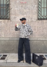 Homme portant une casquette noire, des lunettes de soleil, une veste à motifs et un pantalon large noir, tenant une tasse de café, debout près d’un mur en pierre avec deux fenêtres et un sac noir.