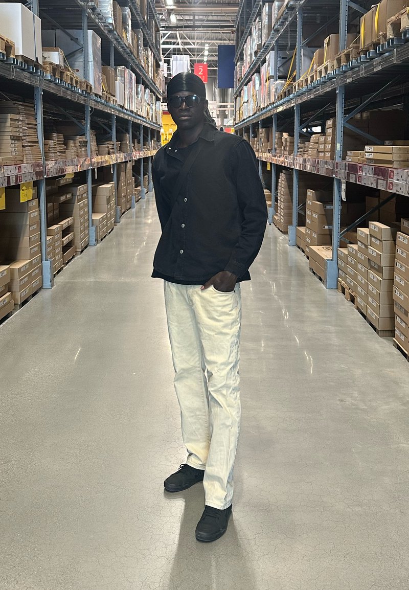 Άνδρας που φοράει μαύρο πουκάμισο, λευκό παντελόνι και γυαλιά ηλίου, στέκεται σε διάδρομο αποθήκης με ψηλά ράφια γεμάτα κουτιά και στις δύο πλευρές.