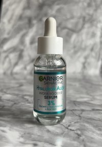 Bouteille compte-gouttes en verre transparent remplie d'un sérum transparent. Capuchon compte-gouttes blanc, étiquettes argentées et vert teal avec les détails du produit, concentration de 3 %.