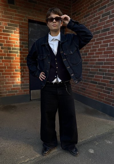 Chaqueta de mezclilla oscura con bolsillos frontales, camisa blanca y chaleco azul marino. Pantalones negros holgados y zapatos negros. La persona lleva gafas de sol y posa al aire libre.