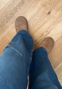 Personne portant un jean bleu et des chaussons en daim marron debout sur un sol en bois clair, faisant face à l'avant.