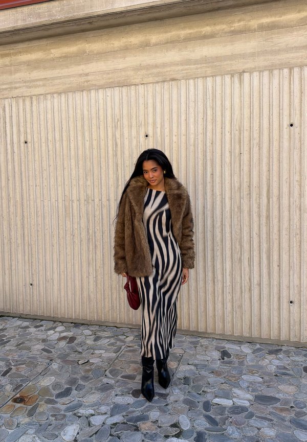 Vrouw draagt een bruine bontjas over een zwart-beige jurk met zebra-strepen, houdt een rode handtas vast en staat op een stenen trottoir tegen een beige muur.