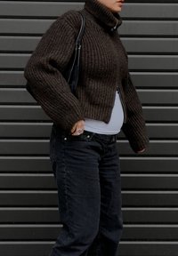 Brun ribbestrikket genser med høy krage og kort liv, kombinert med svarte jeans. En svart veske henger over den ene skulderen.