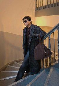 Długi, ciemny płaszcz w cętki na brązowym polo, do tego czarne rękawiczki. Nosi ciemnobrązową, usztywnianą torebkę, schodząc po spiralnych schodach.