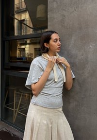 Harmaa lyhythihainen t-paita, kermavärinen neulottu huivi solmittuna kaulan ympärille, vaaleanbeige laskostettu hame, seisoo betoniseinää vasten, sisätilan tausta näkyvissä.