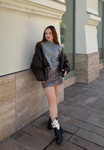 Joven mujer apoyada contra la pared de un edificio, vistiendo un suéter gris, chaqueta negra, falda con estampado y botas en blanco y negro, sosteniendo un teléfono.
