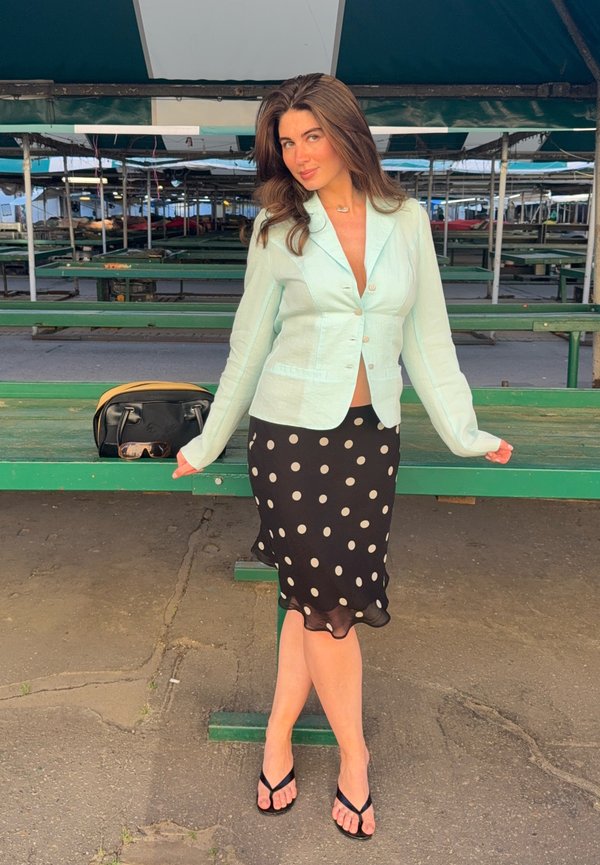 Femme portant un blazer bleu clair et une jupe noire à pois, debout jambes croisées près d'un stand de marché vert sous un auvent.