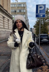 Cappotto bianco oversize in lana con tessuto strutturato; guanti neri; cappello bianco soffice; borsa in pelle marrone; tiene una tazza di caffè e una rivista.