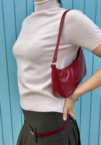 Geantă de mână roșie din piele, cu un design curbat, purtată peste un pulover scurt cu mâneci scurte de culoare roz deschis, asortată cu o fustă plisată de culoare gri închis și o curea roșie.