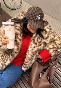 Manteau en fausse fourrure à motif léopard, pull en maille rouge, casquette à carreaux, gobelet isolant rose et sac cabas en cuir marron sur une surface en bois texturée.