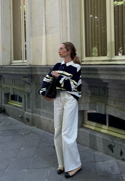 Camisa de rayas oversized en azul marino y blanco con collar, combinada con pantalones anchos blancos y zapatos negros y beige. Sosteniendo un bolso negro.